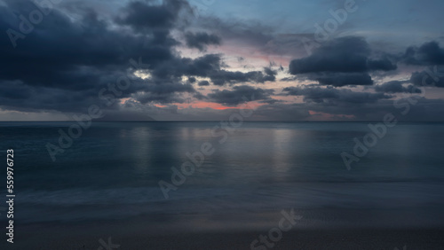 Calm evening seascape in shades of blue. Picturesque clouds in the sky, highlighted in pink. Reflection on the ocean surface. Foam of waves on wet sand. Long exposure. Seychelles. Mahe. Beau Vallon