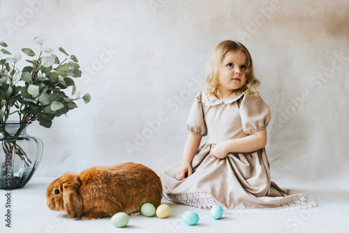 little blonde girl in vintage dress near retro background and vase with green branches eucalyptus gum tree, brown bunny rabbit spring day, happy childhood, simple and cozy life, happy Easter eggs hunt