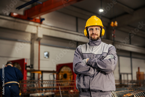 A confident metallurgy worker in protective work wear is standing in facility and smiling at the camera.