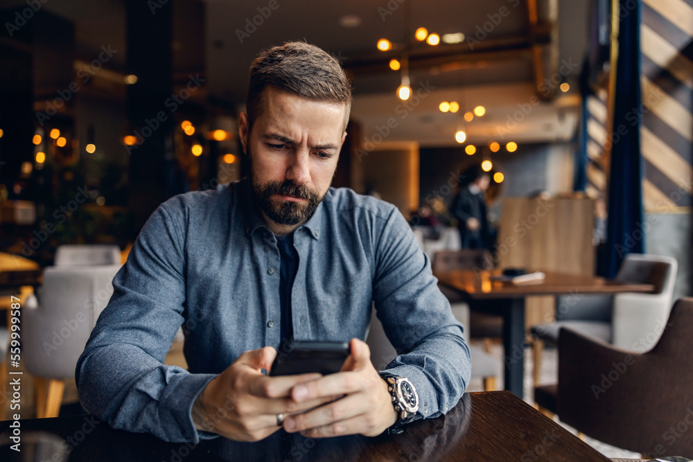 A serious man is sitting in a coffee shop with phone in his hands and reading e-mails.