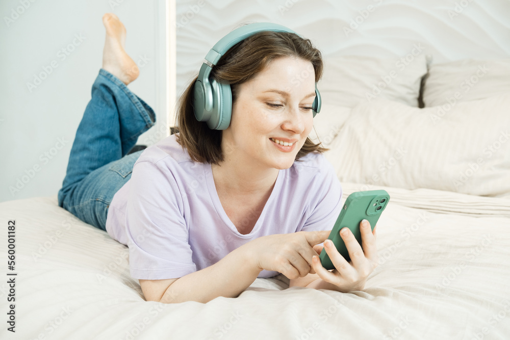 Smiling woman lies on the bed in headphones and listens to music while browsing tracks holding a smartphone in her hands Smiling woman lies on the bed in headphones and listens to music while browsing tracks holding a smartphone in her hands