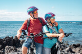 Caucasian senior couple of cyclists wearing helmets resting on beach with electric bicycles enjoying sunny day. Authentic senior retired life concept. Horizon over the sea