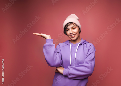 Look here, advertise! Portrait of a happy pretty girl in a lilac sweater holding an empty space on her palms, an empty wall for advertising, a studio shot isolated on a pink background