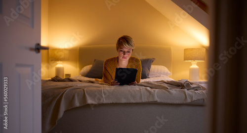 Evening View Through Door Of Young Woman Lying On Bed At Home Looking At Digital Tablet