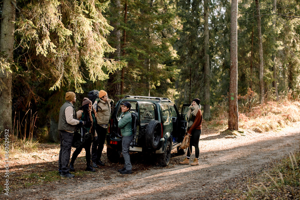 Multiracial male and female friends talking to each other while standing near off-road vehicle on dirt road against tree Multiracial male and female friends talking to each other while standing near off-road vehicle on dirt road against tree