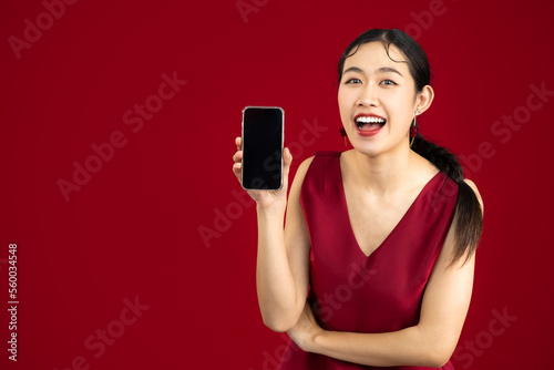 Young beautiful asian woman wear elegant red dress holding and showing blank screen smartphone on red color background.