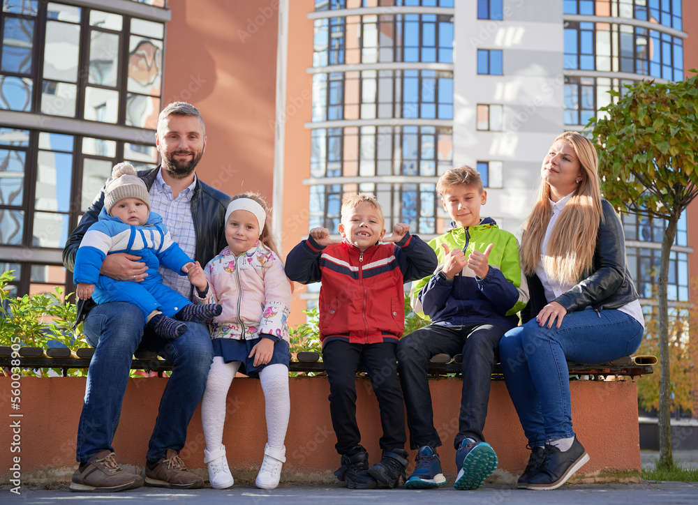 Fototapeta premium Happy family - father, mother and children having fun together on playground. Parents and kids sitting on bench, looking to camera. Modern residential buildings on background.