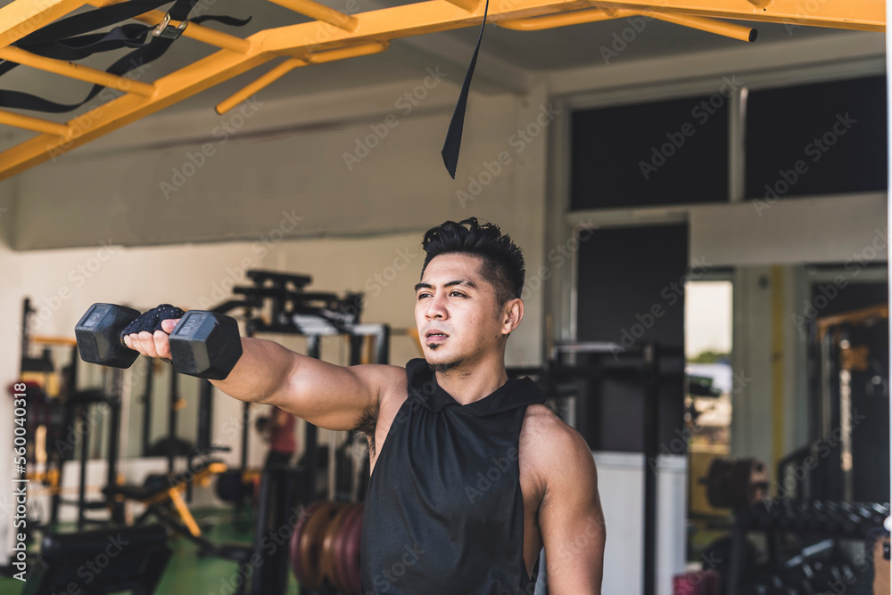 A tanned and handsome asian guy doing standing dumbbell alternating front raises at the gym. Wearing a low cut tank top. Weight training at the gym.