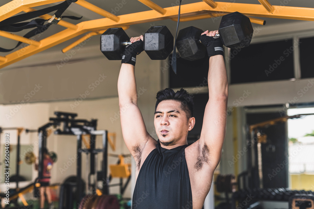 A tanned and handsome asian guy doing standing dumbbell shoulder presses at the gym. Wearing a black hooded low cut tank top. Weight training at the gym. A tanned and handsome asian guy doing standing dumbbell shoulder presses at the gym. Wearing a black hooded low cut tank top. Weight training at the gym.