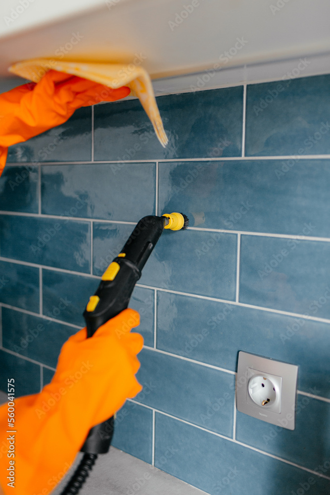 A woman's hand in an orange rubber glove washes the tile in the kitchen using a steam generator. Professional cleaning service. The concept of cleaning the bathroom or kitchen, cleanliness.