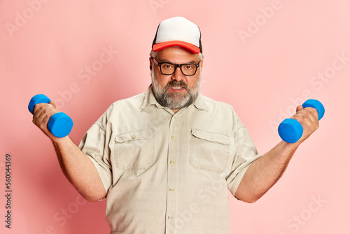 Portrait of mature overweight man training with dumbbells over pink studio background. Sport motivation, body care