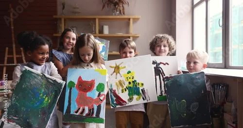 Group of little multi ethnic kids showing their paintings painted during art class smile look at camera. Portrait of happy proud creative children pose together in workshop. Hobby, talent development