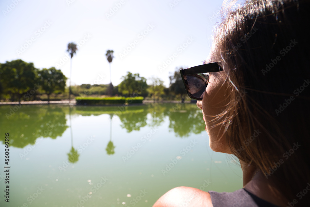 Beautiful, dark-haired Spanish woman is looking at the background where you can see a lake in the park. The woman is dressed in elegant black clothes and wears sunglasses.