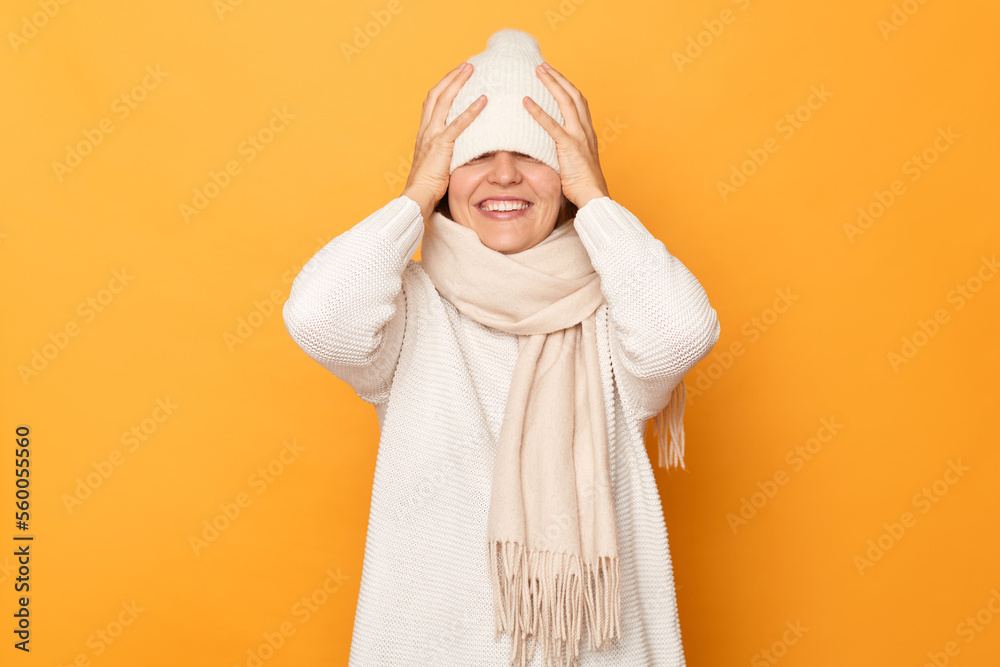 Indoor shot of smiling satisfied woman wearing sweater, scarf and hat, hiding her eyes with cap, smiling happily, having fun, feels shy, standing isolated over yellow background. Indoor shot of smiling satisfied woman wearing sweater, scarf and hat, hiding her eyes with cap, smiling happily, having fun, feels shy, standing isolated over yellow background.