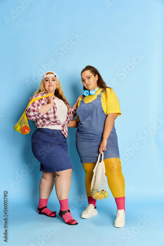 Portrait of two fat, beautiful women, friends posing with bag with fruits and vegetables over blue studio background. Diet