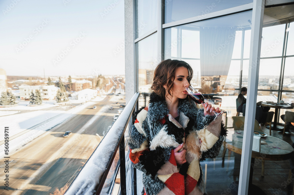 a sexy girl dressed in a long fur coat poses on the balcony against the background of the winter city; holding a glass of red wine