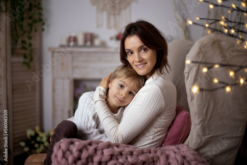 Cute blonde toddler child, boy, cuddling in mother, sitting on an armchair, smiling and hugging