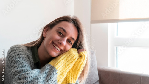 Beautiful woman in winter clothes sitting on sofa at home and looking at camera. Young adult woman with candid smile. International women's day concept for march 8