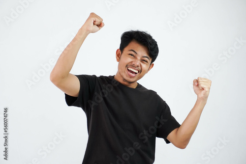 Confident cheerful handsome asian man lifting hands up wearing black t shirt winner gesture clenching fists. feels happiness show fist up success isolated on white background