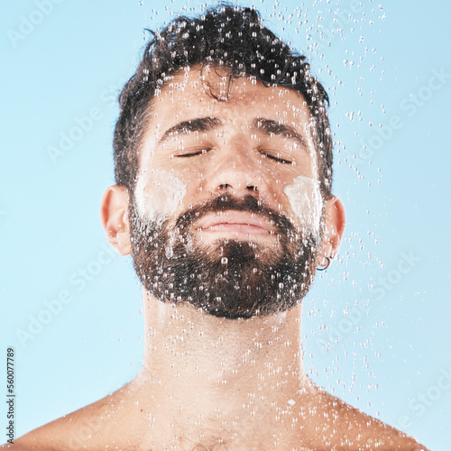 Water, man with face and cream for cleaning in shower with hygiene, grooming and skincare against blue studio background. Clean, model with water drops and facial, natural treatment and cosmetic care