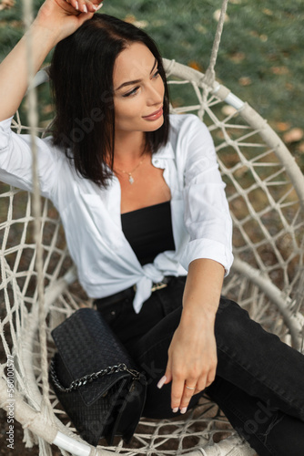 Fashion pretty business woman in stylish elegant outfit with white shirt, black jeans and leather fashion bag sits and rest on knitted swing outdoors 