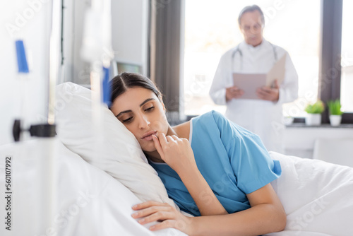 brunette woman in patient gown lying on bed near blurred doctor in clinic.