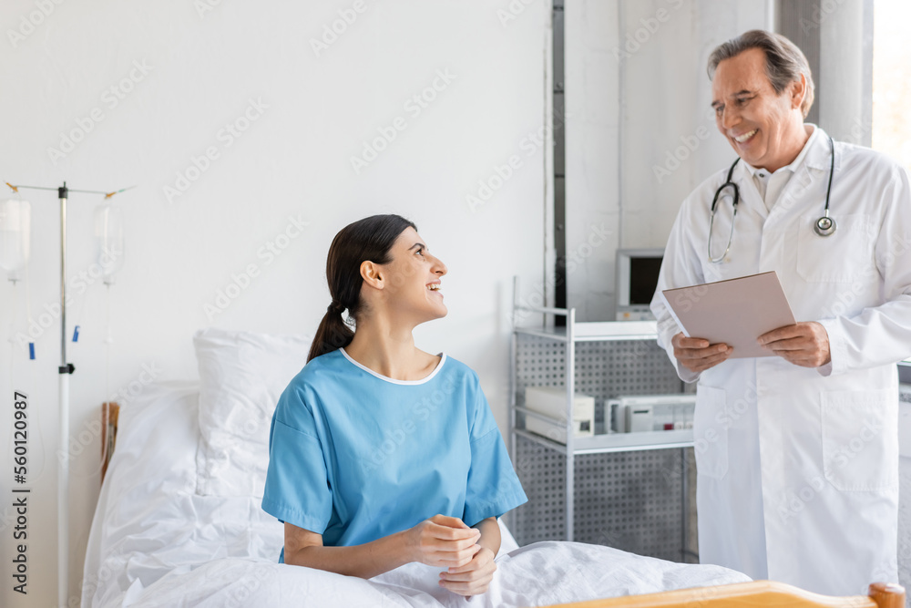 Smiling senior doctor holding paper folder near patient in hospital ward. Smiling senior doctor holding paper folder near patient in hospital ward.
