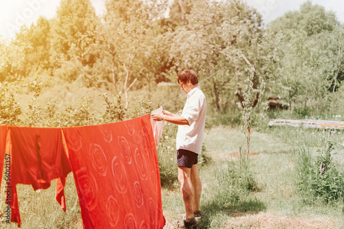 candid young mature choring man chore homework and hanging the laundry to dry on a clothesline on the street in courtyard of village household cottage house. summer, freshness and laundry day. flare photo