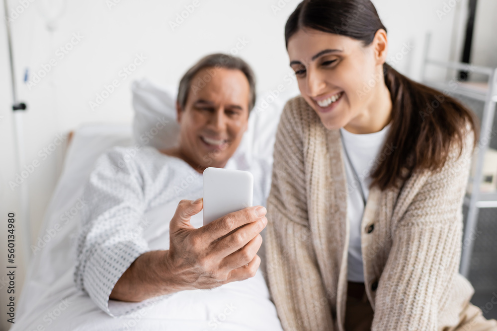 Blurred senior patient holding smartphone near daughter in hospital ward. Blurred senior patient holding smartphone near daughter in hospital ward.