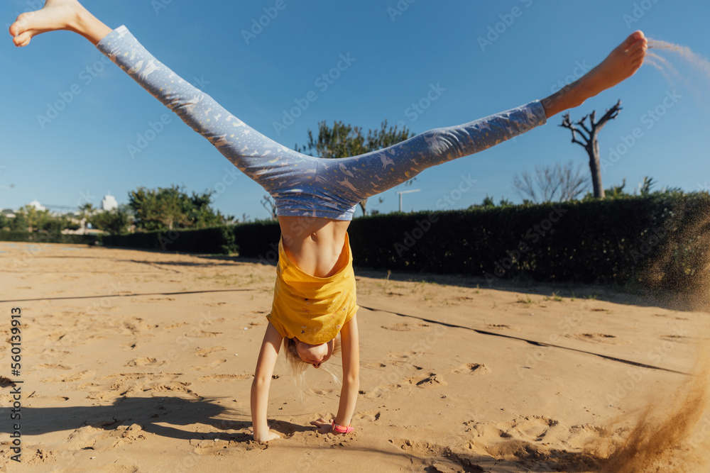 Little girl stands on hands in sand on a sunny day, girl wearing yellow