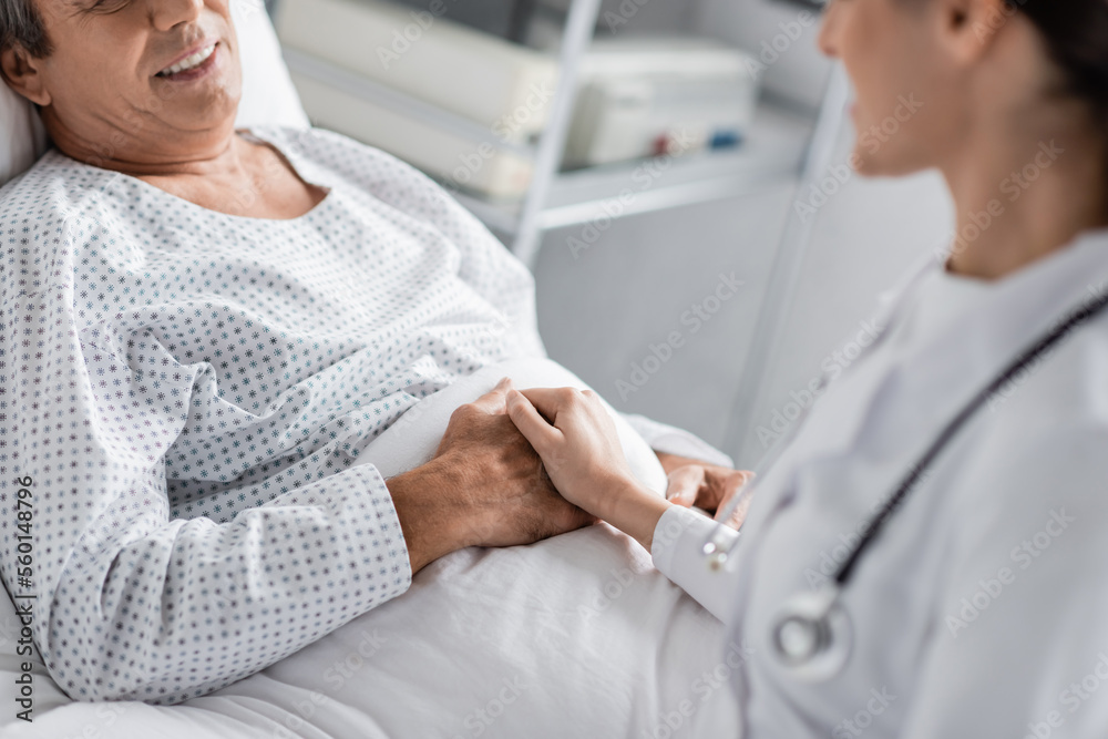 Fototapeta premium Cropped view of blurred doctor holding hand of smiling patient in hospital ward.