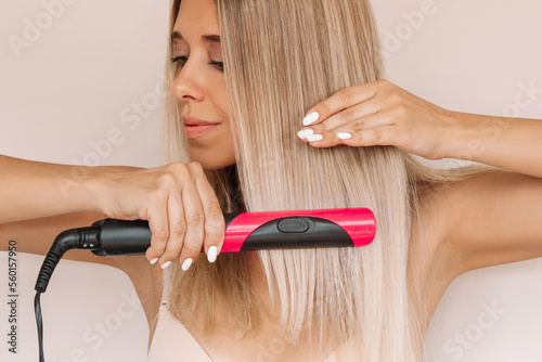 A young attractive caucasian blonde woman straightens her hair with black and pink hair straightener on a light beige background photo