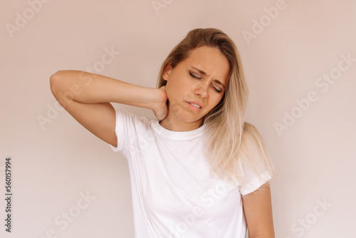 Young attractive caucasian blonde woman in a white t-shirt with a neck pain holding her neck isolated on a beige background. The girl suffers from an osteochondrosis