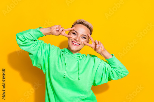Photo of young pretty attractive overjoyed positive girl wear green hoodie sportswear cover face v-sign greetings isolated on yellow color background