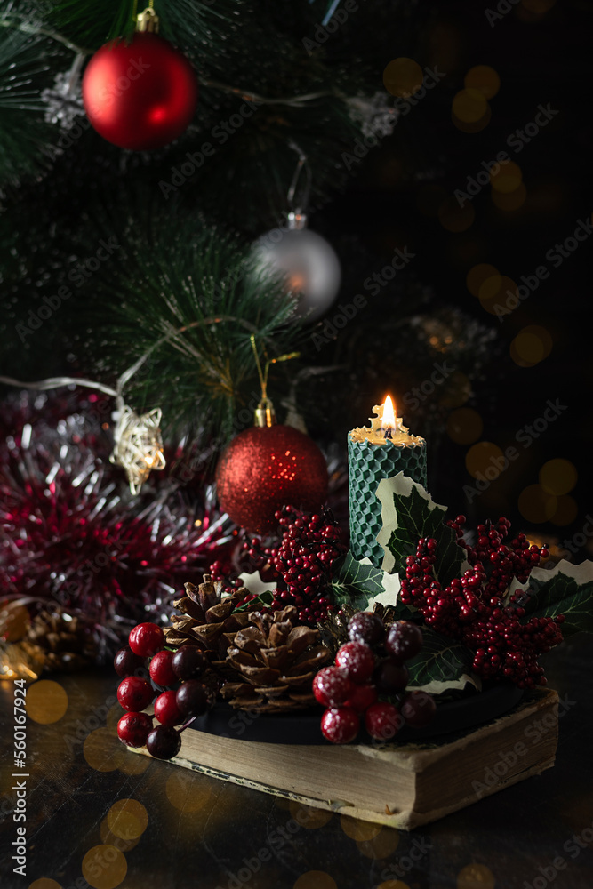  green candle made of natural wax on a dark table. Red berries, fir branches and cones on a black plate. Christmas tree with red balloons and garlands in the background
