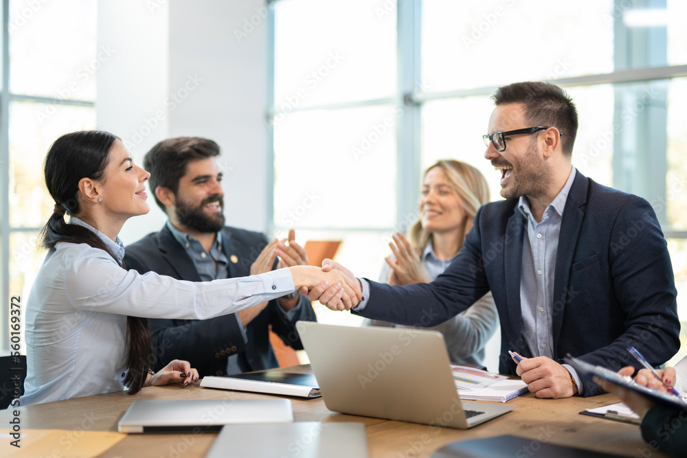 Successful business woman and business man in elegant suit shaking hands together in front of group of business partners clapping hands in modern office. Successful business woman and business man in elegant suit shaking hands together in front of group of business partners clapping hands in modern office.