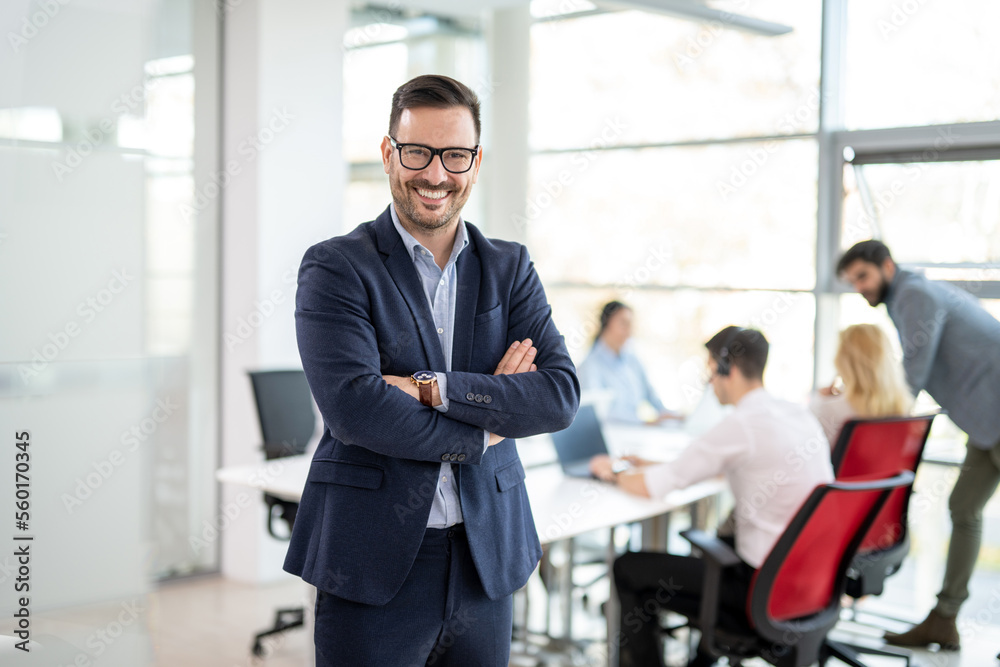 Portrait of confidence business man in formalwear standing with crossed hands at office with colleagues in the background Portrait of confidence business man in formalwear standing with crossed hands at office with colleagues in the background