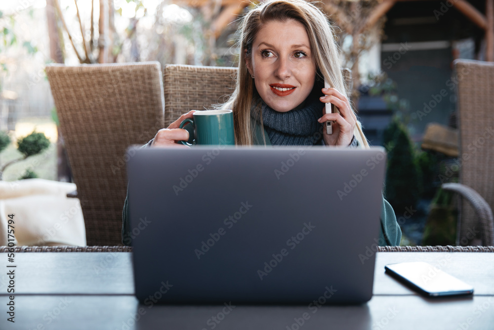 Naklejka premium Beautiful young woman with laptop and talking on cell phone and smiling. Female using cell mobile phone while sitting at home garden outdoors.
