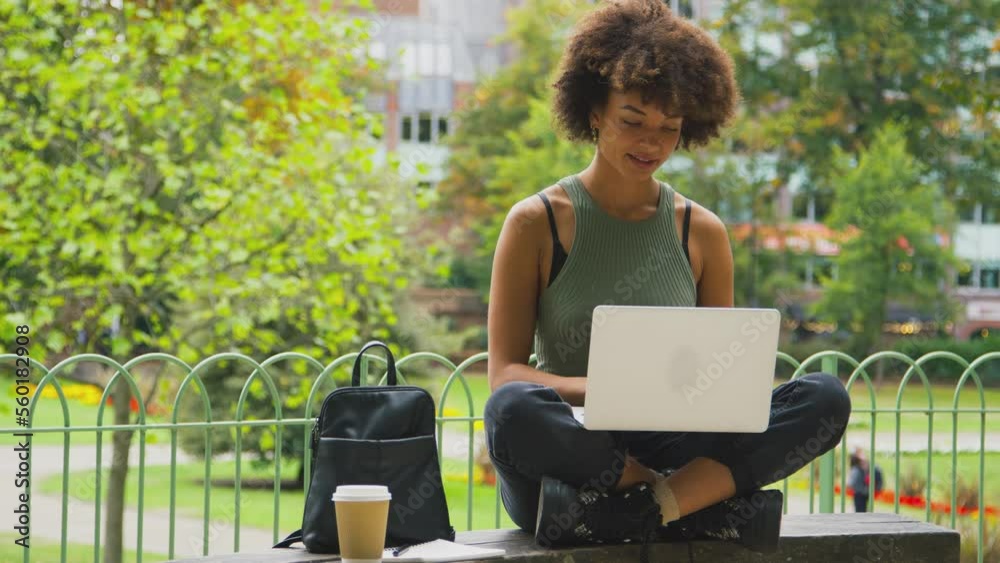 Businesswoman sitting on bench in city park working on laptop with takeaway drink - shot in slow motion Businesswoman sitting on bench in city park working on laptop with takeaway drink - shot in slow motion