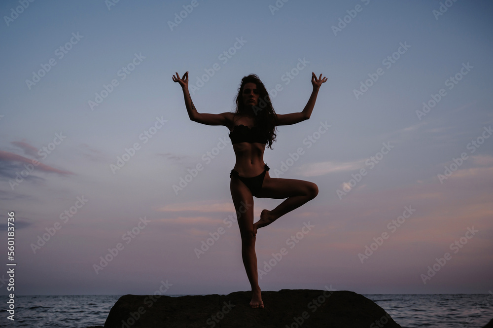 Silhouette of beautiful young girl in black swimsuit standing in a yoga pose on the beach by sea in summer