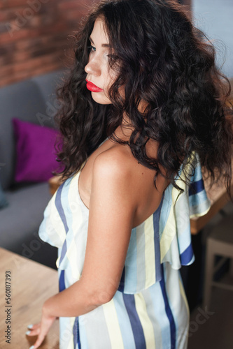 curly sexy brunette poses in the cafe hall; wearing a light dress; lips painted with red lipstick