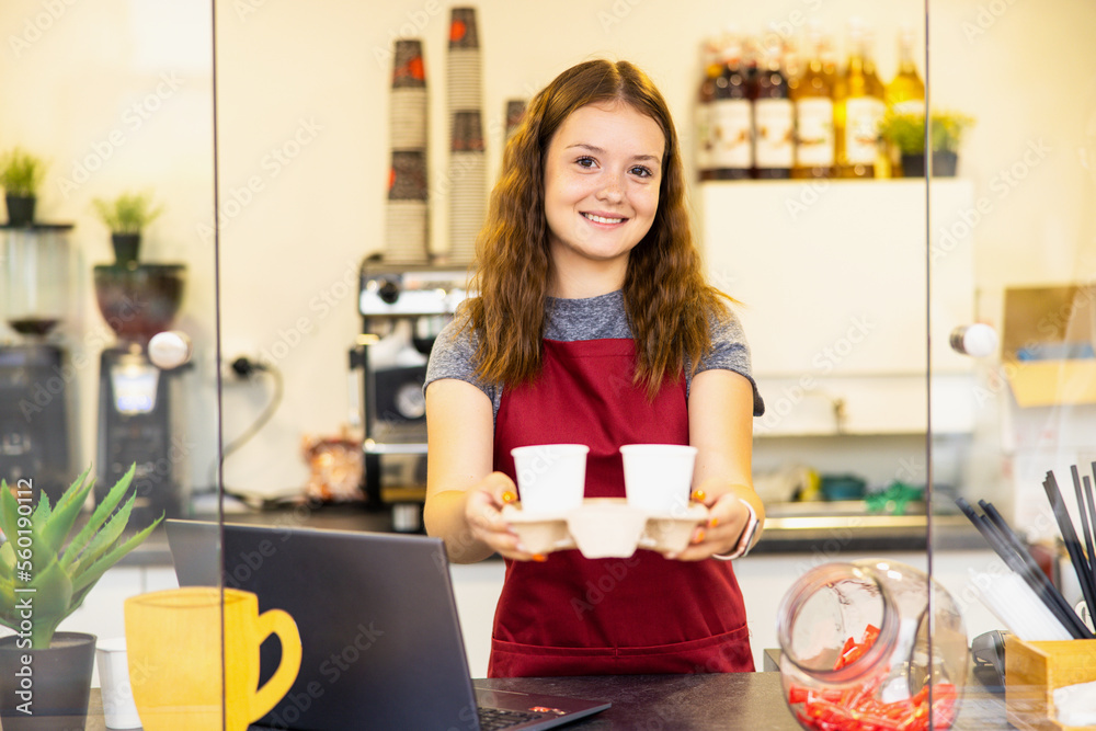 Portrait of a young beautiful woman in red apron, keeps white cup in hand, smiling, looking at camera, on a coffee shop background. Portrait of a young beautiful woman in red apron, keeps white cup in hand, smiling, looking at camera, on a coffee shop background.