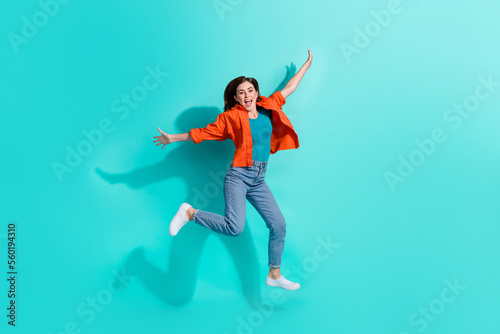 Full length photo of funny excited girl dressed orange shirt jumping high arms sides isolated turquoise color background