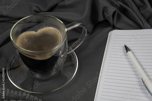 Herzförmige Glastasse mit Kaffee gefüllt auf Glasunterteller vor grauem Satinstoff, ein weißer Kugelschreiber auf liniertem Notizheft rechts noch etwas mit im Bild, horizontal photo