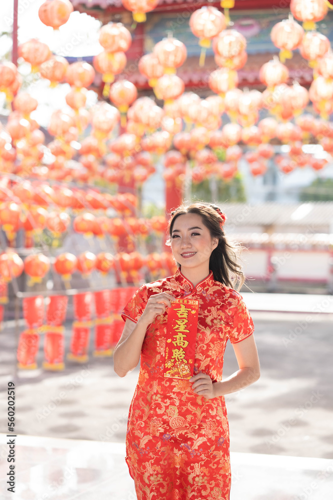 Obraz premium Happy Chinese new year. Young asian woman wearing traditional cheongsam qipao dress holding blessing fortune card in Chinese Buddhist temple. Chinese text means blessed by a lucky star. Emotion Smile