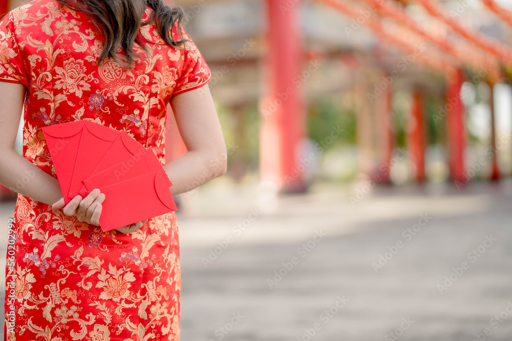 Happy Chinese new year. Beautiful asian woman wearing traditional cheongsam qipao dress and stands with her back turned holding ang pao, red envelopes in Chinese Buddhist temple.