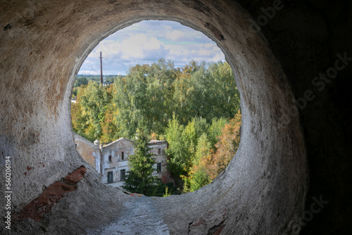 A large round hole in the shabby brick wall. View through a cement pipe to the countryside. In the distance  a blurred landscape  forest  cloudy sky  outlines of buildings. 