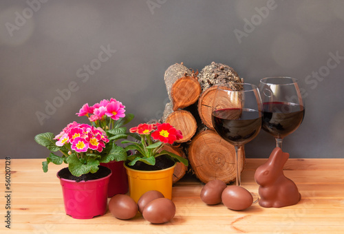 Easter concept. Primula flowers, glasses of red wine, chocolate easter eggs, chocolate bunny and  pile of firewood on wooden table.