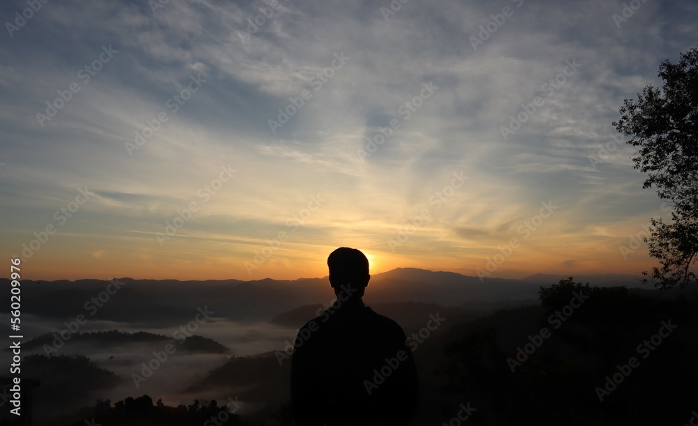 A man facing in front of the sun is rising from a back mountain and there has a sea of fog too.