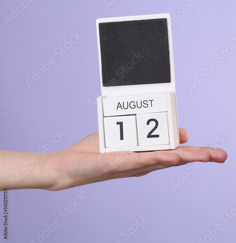 Woman's hand holds wooden calendar with date august 12 on pastel purple background